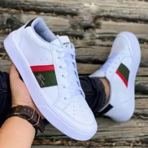 Tenis Lacoste blancos de diseño urbano con franjas roja y verde, logo metálico lateral y suela de caucho, sostenidos en mano sobre fondo de madera.